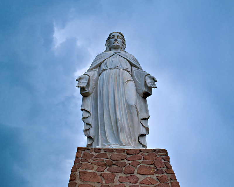 Christ of the Wichitas Overlooks the Refuge White stone statue of Jesus with arms outstretched on red rock pedestal under cloudy sky.