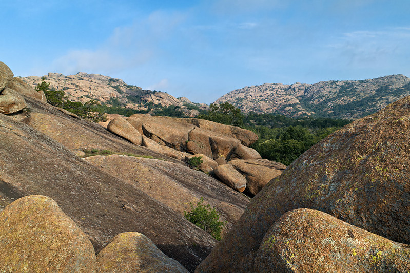 Expansive view of granite boulders and hills with distant mountains and green vegetation in the Wichita Mountains Wildlife Refuge.