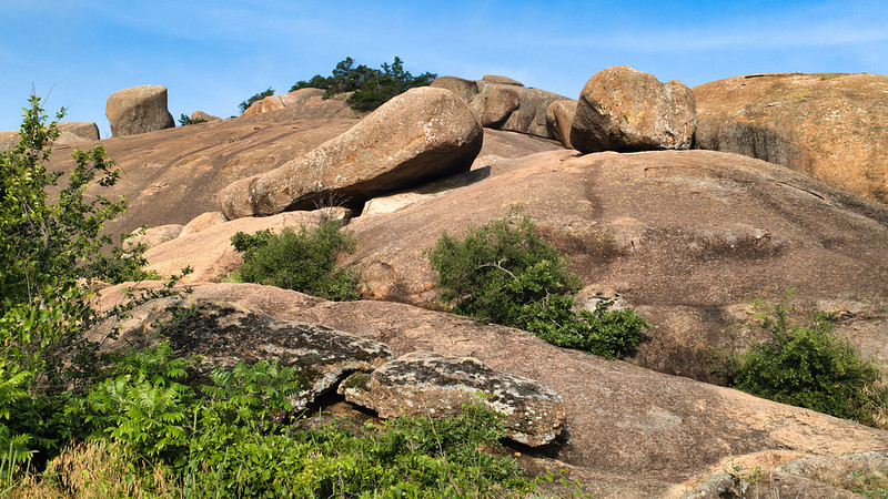 Perched Boulders on a Granite Slope Large granite boulders perched on a slope with green shrubs and blue sky in the Wichita Mountains Wildlife Refuge.