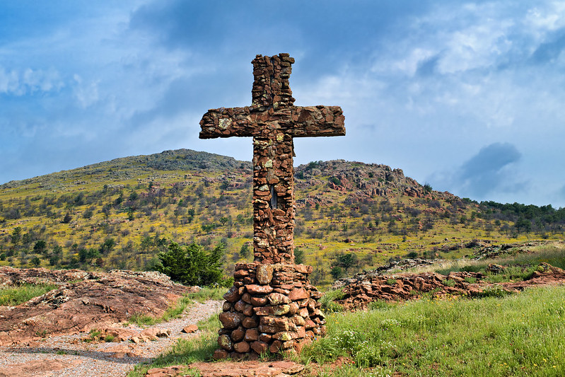 Stone Cross in the Red Hills Rustic stone cross set against red dirt and grassy terrain with scattered wildflowers.