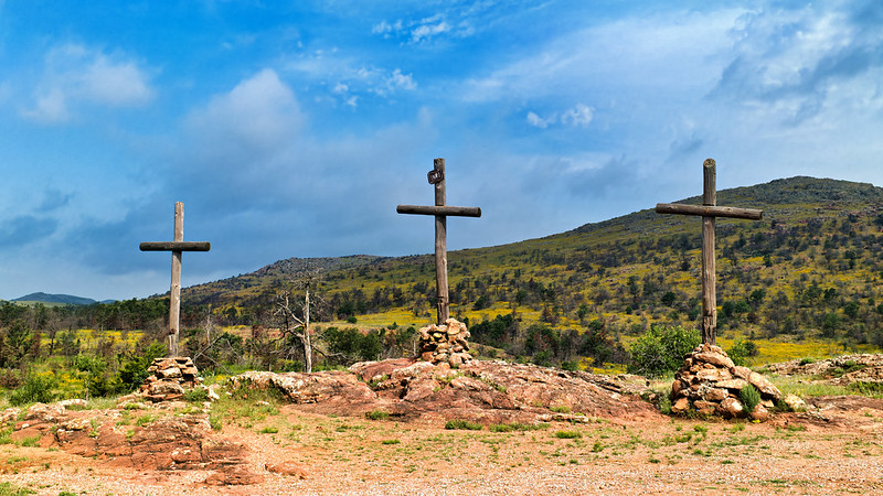 Three Crosses Above the Prairie Three wooden crosses on a rocky platform with grassy hills and cloudy sky in background.